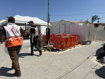 To menn med vester frå Raudekrossen og Raude Halvmåne går på ein steinlagt veg mellom kvite telt og oransje feltsenger i ein leir.