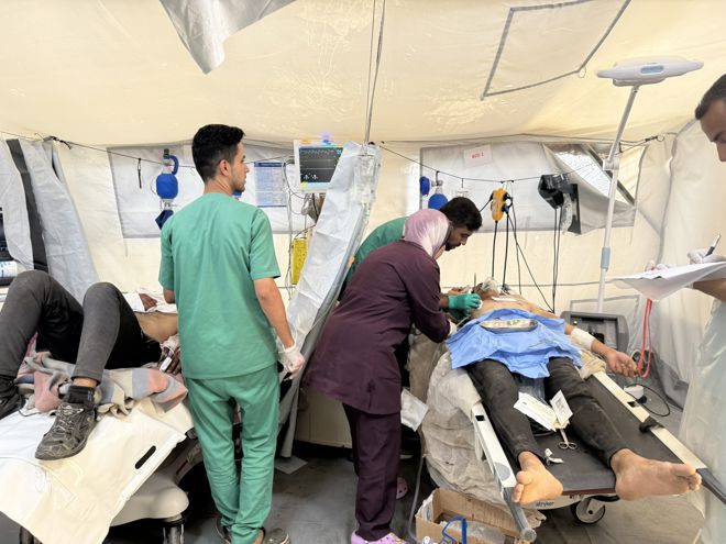 Medical personnel in scrubs treat patients lying on beds inside a temporary medical tent.