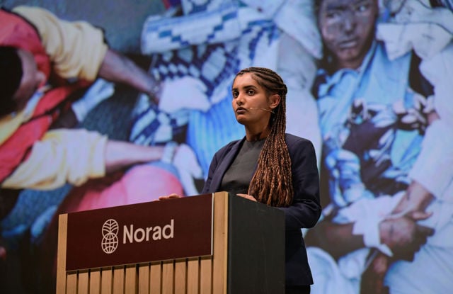 En ung kvinne står bak en talerstol merket med Norad-logoen, foran en stor skjerm som viser et bilde av mennesker i bakgrunnen.