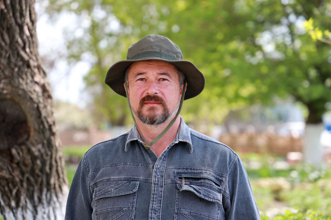 An adult man wearing a wide-brimmed hat and a denim shirt stands outdoors in a green, leafy area.