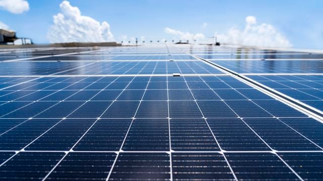 Bildet viser en nærbilde av solcellepaneler som strekker seg horisontalt over en flate. Panelet har en blank overflate som reflekterer solens lys. I bakgrunnen kan man se en klar blå himmel med noen spredte, hvite skyer.