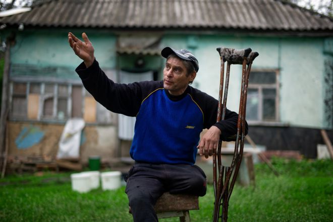 A man with one leg, wearing a blue and black sweater and a cap, sits on a stool in front of an old house, holding a pair of crutches with one hand and gesturing with his other arm raised.