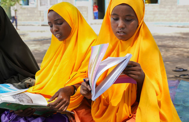 Two girls in yellow hijabs sit outdoors reading schoolbooks.