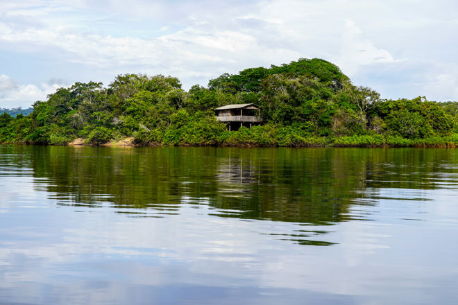 Bildet viser en frodig, grønn øy med tett vegetasjon. Midt på øya står et enkelt, trekledd hus på påler, delvis skjult av trærne. Øya er omgitt av stille vann som speiler både hus og vegetasjon. Himmelet er lys med noen skyer, og landskapet gir en følelse av ro og isolasjon, langt fra by eller tettsted.