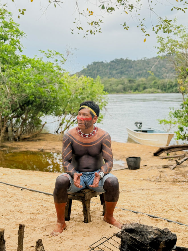 En mann sitter på en enkel trestol på en sandstrand ved elvebredden. Han har omfattende kroppsmaling i sorte og røde mønstre over overkroppen, armene og bena