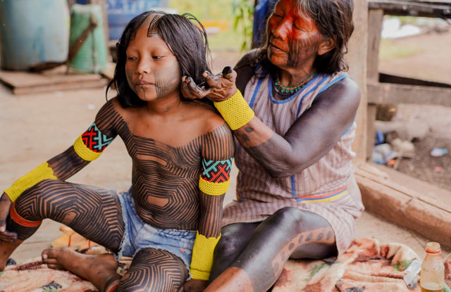 The image shows two people, an adult woman and a child, sitting on a blanket outdoors. The woman is holding and tending to the child's hair. Both have their bodies adorned with extensive black patterns that resemble traditional body painting.