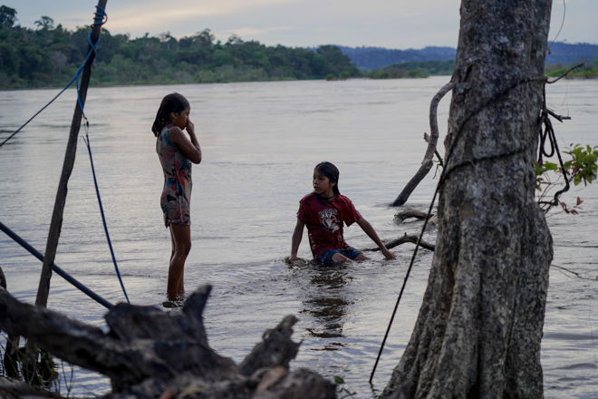 Bildet viser to barn ved elvebredden i et naturlandskap. Den ene står i vannet med en kort sommerkjole, mens den andre sitter på huk i vannet med en rød T-skjorte og shorts. Vannet er rolig, og det er trær og vegetasjon rundt dem, med en skog i bakgrunnen.