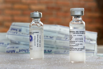 Bildet viser to små glassflasker med gjennomsiktig væske, plassert på et bord med is eller et kjøleelement. Flaskene har etiketter som indikerer at de inneholder vaksine mot difteri og stivkrampe.