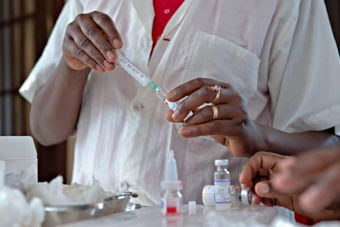 En person iført hvit skjorte trekker opp væske fra et lite glass medisinflaske inn i en sprøyte. På bordet foran ligger bomullsdotter, flere små medisinflasker og utstyr. 