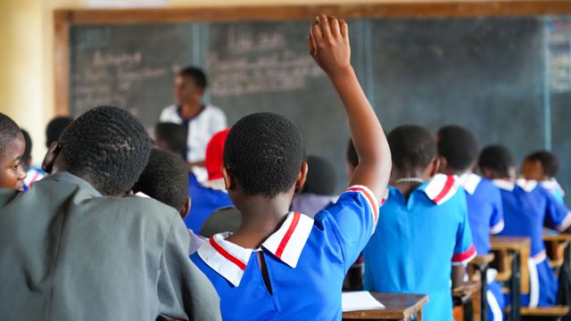 Elev i blå skuleuniform rekkjer opp handa i klasserom med fleire elevar framfor tavle.