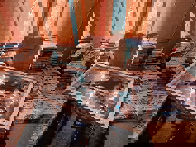 The image shows an office space with stacks of newspapers piled on desks and tables. Some of the bundles are labeled with years such as "2022" and "2023." In the center, there is a large chair and a broken window frame with shattered glass resting on it. The background features orange vertical blinds, and the overall setting looks cluttered and somewhat chaotic.