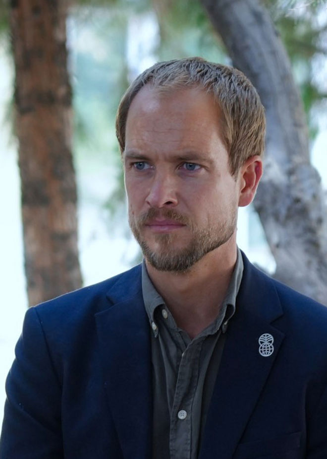 Portrait of Erik Abild. He stands with short blond hair wearing a shirt and dark jacket with a Norad logo pin, outdoors in front of trees.