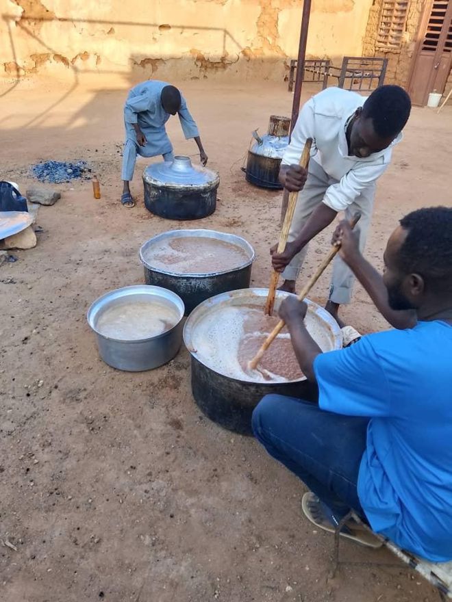Three men are outdoors stirring large pots of food over open fires on the ground, with several metal containers nearby.