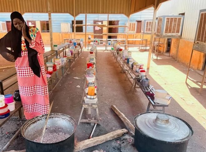 The image shows a woman wearing a long red-and-white patterned dress and a black headscarf standing by a large pot over an open fire. The pot is on the ground, and there is a large wooden ladle inside it. Around her are benches and tables with many colorful plastic containers and pots, likely filled with food or ingredients.