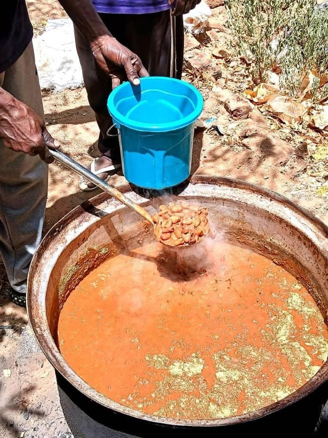 The image shows a large pot of reddish-brown stew or soup boiling outdoors. One person is holding a ladle with beans or meat over the pot, and another person is holding a blue plastic bucket ready to receive food.