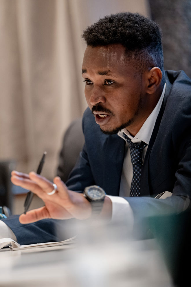 A young man from Sudan sits at a table wearing a suit and tie. He holds a pen and has his hand raised, as if actively taking part in a conversation or meeting. In front of him on the table lies an open sheet of notes.