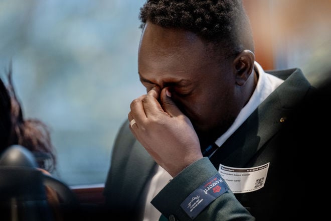 The image shows a man from Sudan, dressed in a dark suit jacket and a white shirt. He is sitting indoors and holding his hand up to his face.
