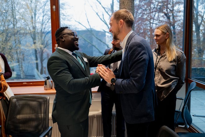 Two men stand close to each other in the middle of the room, greeting or chatting with a handshake and light physical contact, both dressed in suits.