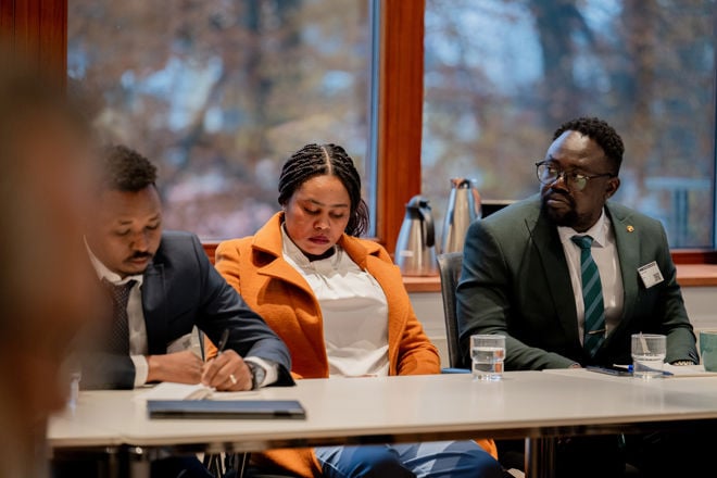 The image shows three people sitting at a meeting table in a bright room. Two men are dressed in suits and ties, and a woman sits between them wearing an orange coat over a white top. There are glasses of water on the table in front of them, as well as notebooks and pens.