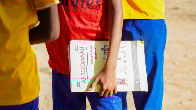 Bildet viser flere barn iført fargerike skoleuniformer. En av dem holder en bok med teksten "Af-Soomaali" på omslaget. Barna har på seg røde og gule t-skjorter med blå bukser, og de står på en utendørs sanddekket område.