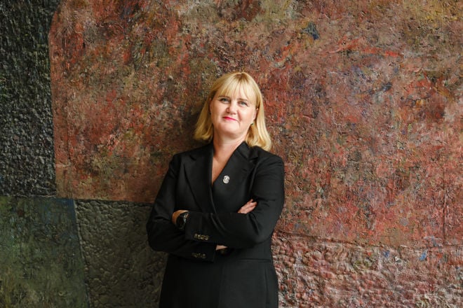 En kvinne med lyst, skulderlangt hår står foran en fargerik vegg iført en mørk dressjakke med en brosje.