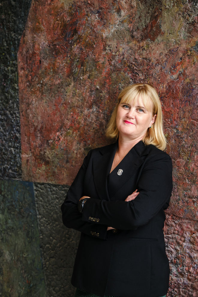 Bildet viser en voksen kvinne med skulderlangt lyst hår som står med armene i kors. Vedkommende har på seg en mørk dressjakke med en brosje på jakkeslaget.