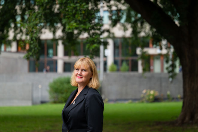 En kvinne med lyst hår og mørk dressjakke står ute på en plen foran en bygning og et stort tre.