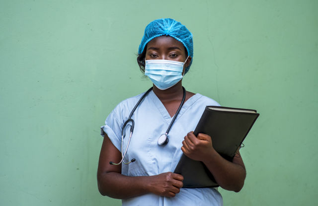 Kvinne i sykehusuniform står med en mappe foran en grønn vegg. 