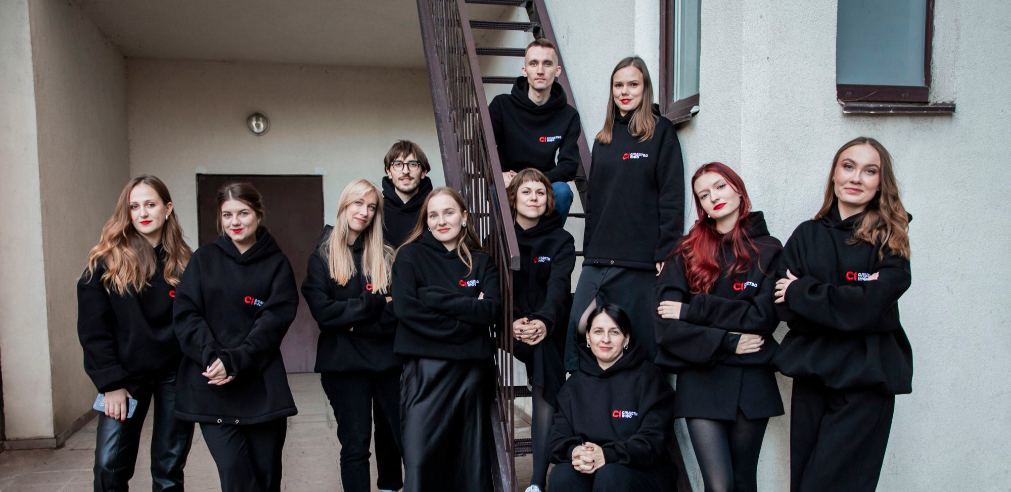 A group of young women are standing and sitting together in front of an outdoor staircase, all wearing black hoodies with a logo on the chest.
