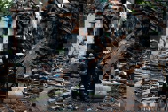 En kvinne med pressevest står og fotograferer ødelagte bygninger og ruiner utendørs.