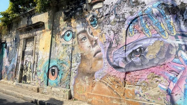 Bildet viser en slitt murvegg utendørs, dekket med fargerik gatekunst. Midt på veggen er det malt et realistisk ansikt av en ung person som ser oppover, omgitt av store, markerte øyne i ulike farger. Til høyre i bildet ser man en stor hånd med blå og gule fingre som holder en slange, der slangens hode har et intenst blikk. Veggen er ujevn, med avskalling og sprekker som gir maleriene et røft preg. Sollys og noen skygger fra trær kan sees øverst på veggen. Stemningen er livlig og fantasifull.