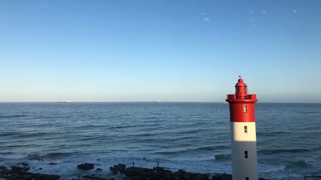 og denne Bildet viser en vakker kystscene med en rød og hvit fyrlykt som står til høyre i forgrunnen. Fyret er slankt og høyt, og det skinner i sollyset. Bakgrunnen består av en uendelig havflate som strekker seg mot horisonten, hvor det er noen få skip som kan sees langt i det fjerne.