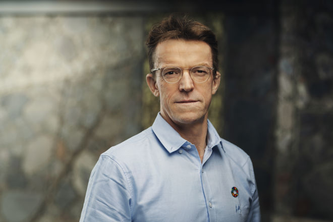 En mann med brunt hår iført lyseblå skjorte med bærekraftsmål-pin, står foran en steinvegg.