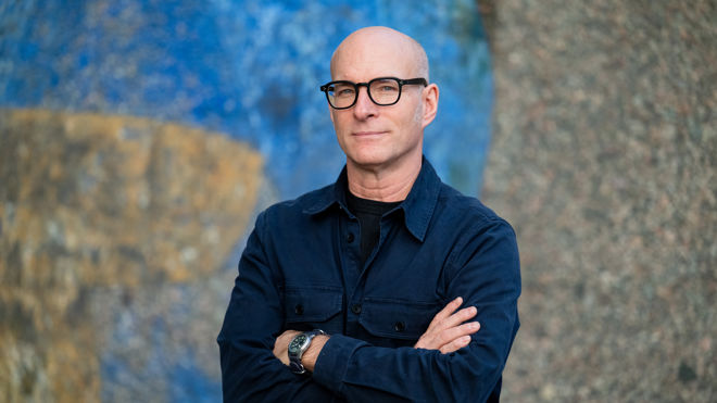 En voksen mann med briller og mørk jakke står med armene i kors foran en blå og grå bakgrunn.