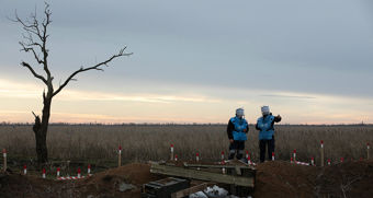 Dette bildet viser et åpent landskap under en grå og overskyet himmel, med et svakt lys fra soloppgang eller solnedgang i horisonten. I forgrunnen står et enslig, bladløst tre med krokete grener, noe som gir scenen et øde og litt dystert preg. To personer står rett ved siden av hverandre, kledd i blå beskyttelsesvester og hjelmer som dekker hele hodet. De ser ut til å være i arbeid med noe farlig, for eksempel minerydding.