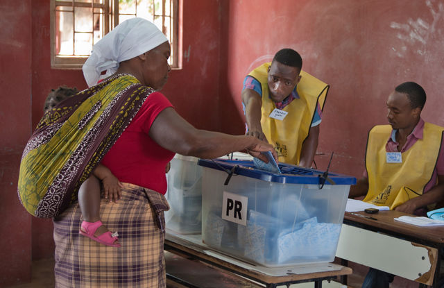 En kvinne med et lite barn på ryggen legger en stemmeseddel i en gjennomsiktig valgurne mens to valgfunksjonærer sitter ved siden av.