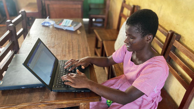 Bildet viser en kvinne som sitter ved et trebord med øynene lukket og jobber på en bærbar datamaskin. Personen er kledd i en rosa- og hvitstripet kjole. På bordet ligger det også en annen lukket bærbar datamaskin og noen bøker eller hefter. 