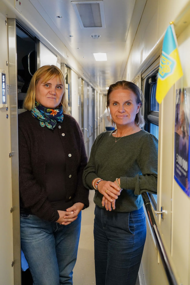 To kvinner står i en smal togvogn og ser i kameraet. 