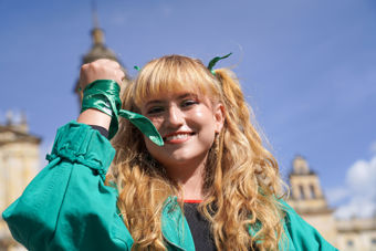 Bildet viser en kvinne med langt, bølgete blond hår, kledd i en grønn jakke. Vedkommende står ute, med en klar blå himmel og noen historiske bygninger i bakgrunnen. Rundt håndleddet og håret har personen knyttet grønne bånd, og holder hånden opp mot ansiktet i en gest som kan tolkes som styrke eller engasjement.