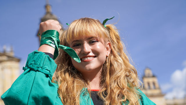 Bildet viser en smilende ung kvinne med langt, bølget blondt hår som er kledd i en grønn jakke. Personen har også grønne bånd knyttet rundt håndleddet og håret, som gir en følelse av koordinerte farger. Bakgrunnen består av en klar blå himmel og en bygning, muligvis en historisk konstruksjon med tårn og detaljer.