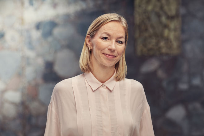 Ei kvinne med lyst, skulderlangt hår står framfor ein uklar bakgrunn og har på seg ein lys skjorte med krage.