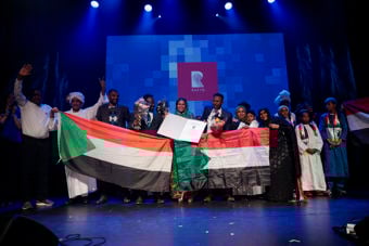 The photo shows a group of people on a stage holding large Sudanese flags, flowers, and a certificate, with a bright blue backdrop and the Rafto logo behind them. They stand close together and smile toward the camera under strong stage lights.