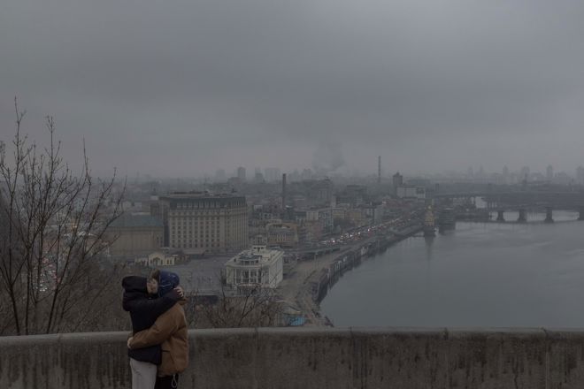 To personer klemmer hverandre ved et rekkverk med utsikt over en stor by og en elv på en grå og overskyet dag.