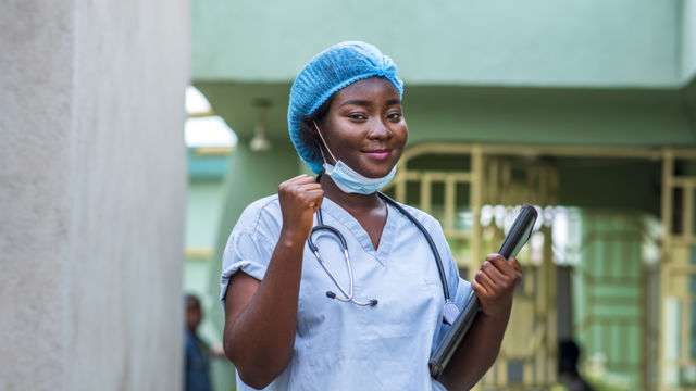 Bildet viser en person i medisinsk bekledning, inkludert en lyseblå skrubbe og en blå hårhette. Personen har en stetoskop rundt halsen og holder en mappe eller dokument under armen. 