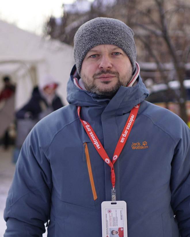 En voksen mann med blå jakke, grå lue og Røde Kors-ID-kort henger rundt halsen, står ute foran et telt i vintermiljø.