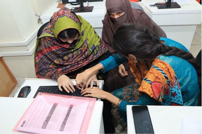 Bildet viser tre kvinner som samarbeider ved et bord med en datamaskin. To av dem peker og jobber sammen på tastaturet. Den ene har en fargerik grønn sjal med mønster, mens den andre har på seg en blå og oransje drakt med et mønstret design. På bordet ligger også en rosa mappe med et dokument inni, og en mobiltelefon. De ser ut til å være engasjert i en felles oppgave i et moderne innendørs miljø, muligens et klasserom eller et kontor.