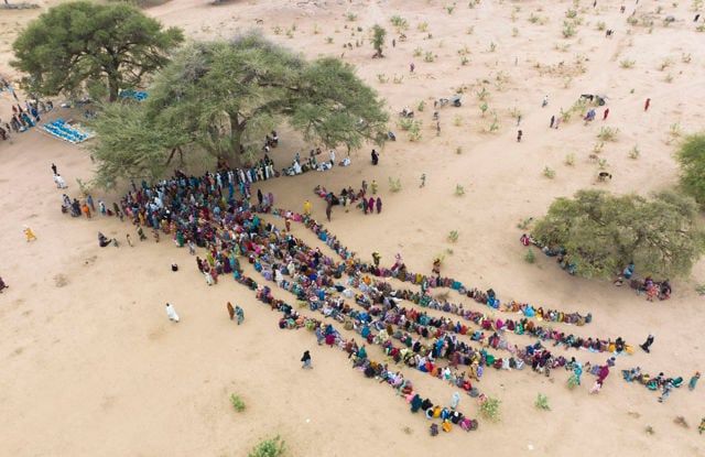 Dette bildet viser en stor gruppe mennesker samlet i et åpent, tørt område. De fleste av dem sitter i lange, parallelle rekker som strekker seg ut på bakken. Mange av dem har fargerike klær som skaper et levende mønster mot den lyse sanden.