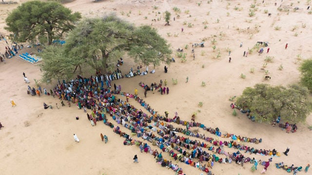 Dette bildet viser en stor gruppe mennesker samlet i et åpent, tørt område. De fleste av dem sitter i lange, parallelle rekker som strekker seg ut på bakken. Mange av dem har fargerike klær som skaper et levende mønster mot den lyse sanden.