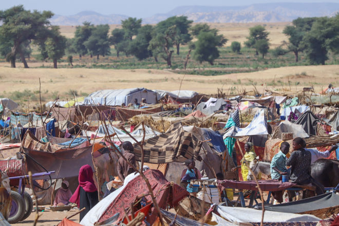A temporary refugee camp with makeshift shelters made of fabric and sticks, where women and children are gathered in a dry, open landscape with scattered trees in the background.