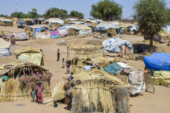 Several simple huts made of straw and tarpaulins stand close together in a dry landscape, with adults and children moving between the huts.